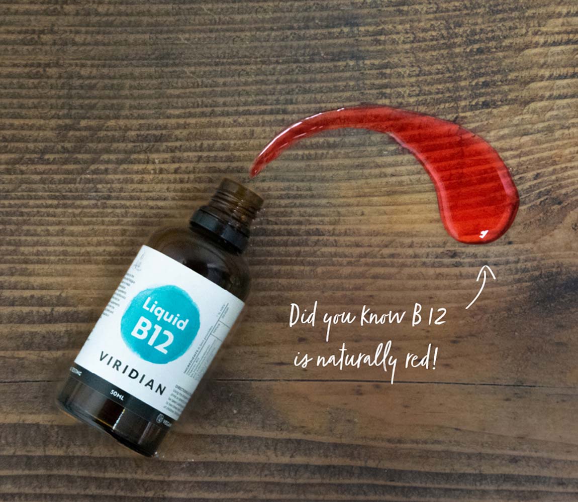 A bottle of VIRIDIAN Liquid B12 supplement lies on a dark wood surface next to a prominent red liquid smear, with handwritten text noting 'Did you know B12 is naturally red!'.