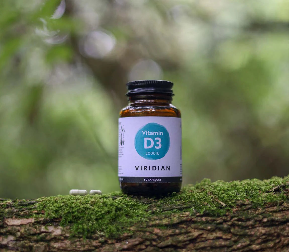 An amber glass bottle of VIRIDIAN Vitamin D3 20000 IU capsules sits on a mossy log outdoors with a soft, blurred green forest background.
