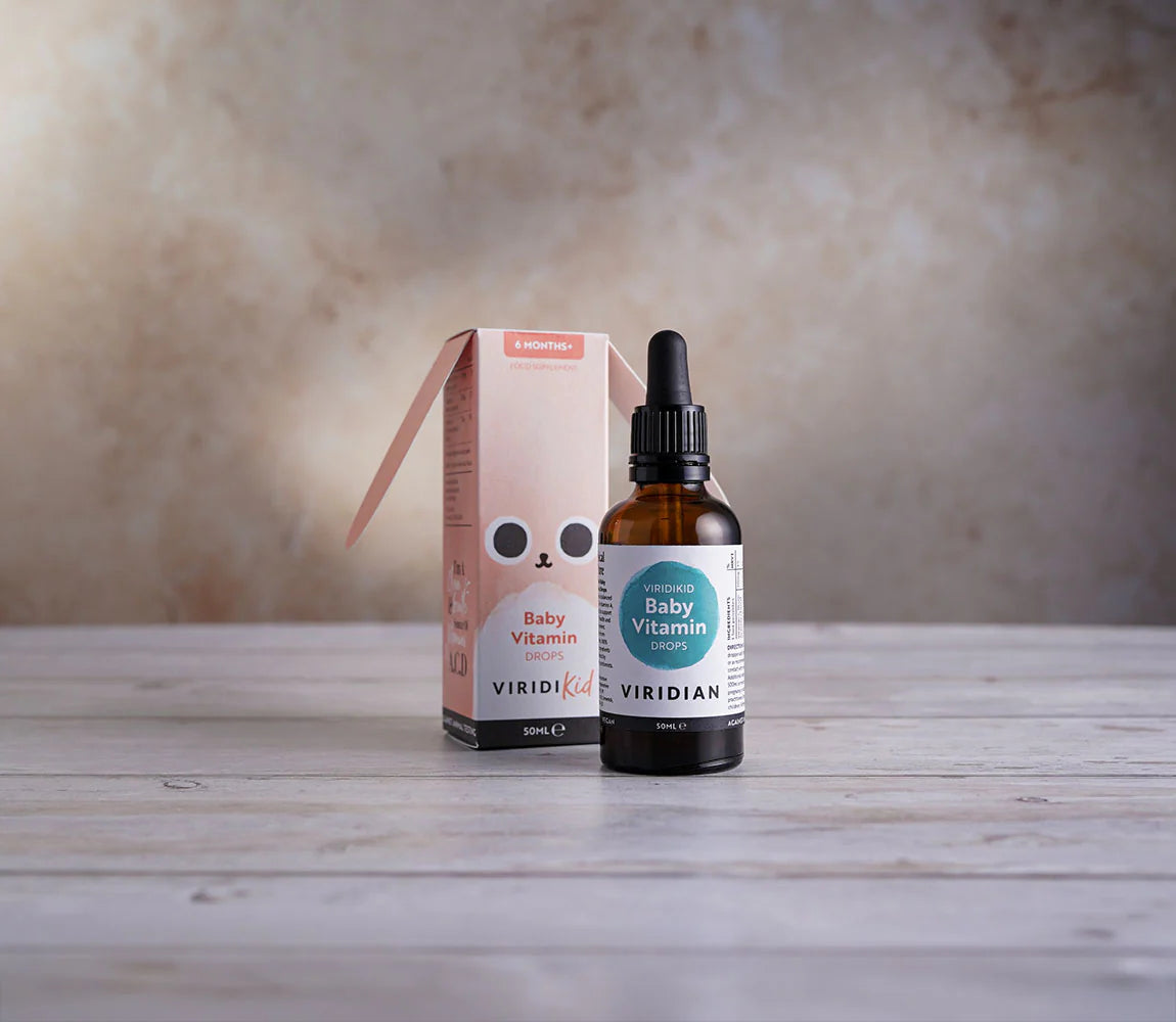 A bottle of Viridian Organic Baby Vitamin Drops next to its pink and white cardboard packaging box, displayed on a light wooden surface against a textured beige background.