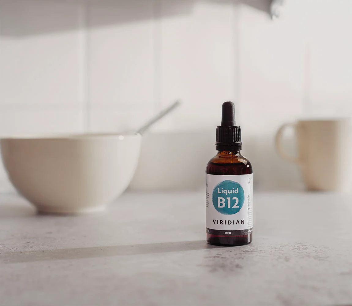A small amber glass dropper bottle labeled 'Liquid B12 VIRIDIAN' stands on a light countertop in a softly blurred kitchen setting with a bowl and mug in the background.