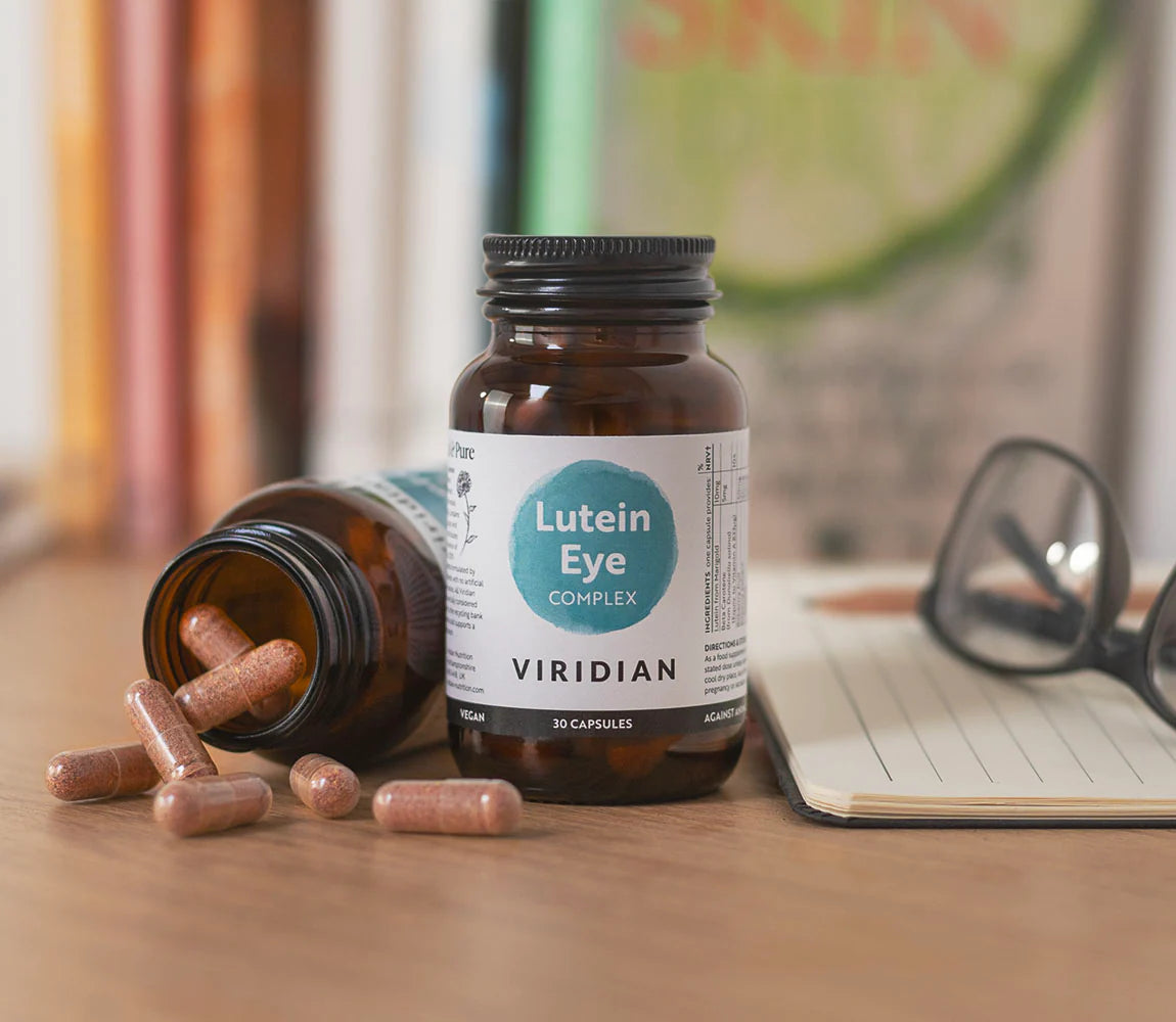 A bottle of Viridian Lutein Eye Complex capsules stands on a wooden table next to an overturned bottle with spilled capsules, with eyeglasses and an open notebook in the background.