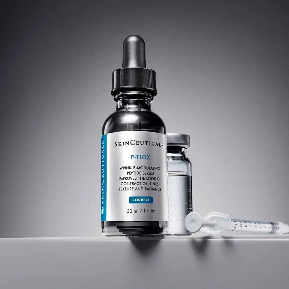 A glass dropper bottle of SKINCeuticals P-TIOX serum stands on a white surface against a dark gray gradient background, with a small vial and a syringe placed behind it.