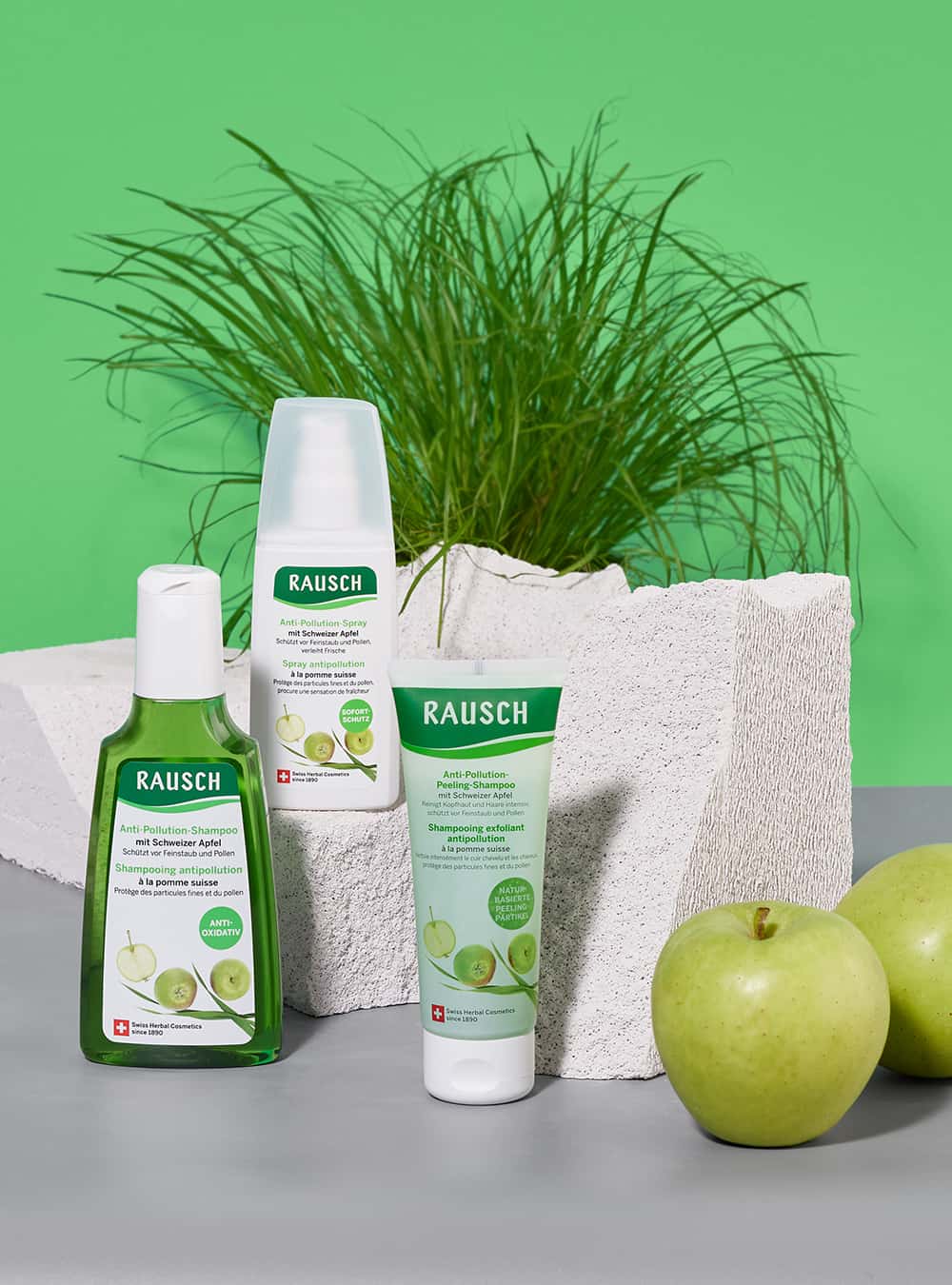 Three Rausch oral care products arranged on a gray surface with a green backdrop and a white textured planter: a green bottle, a white tube, and a white bottle, with green apples in front and a plant behind.