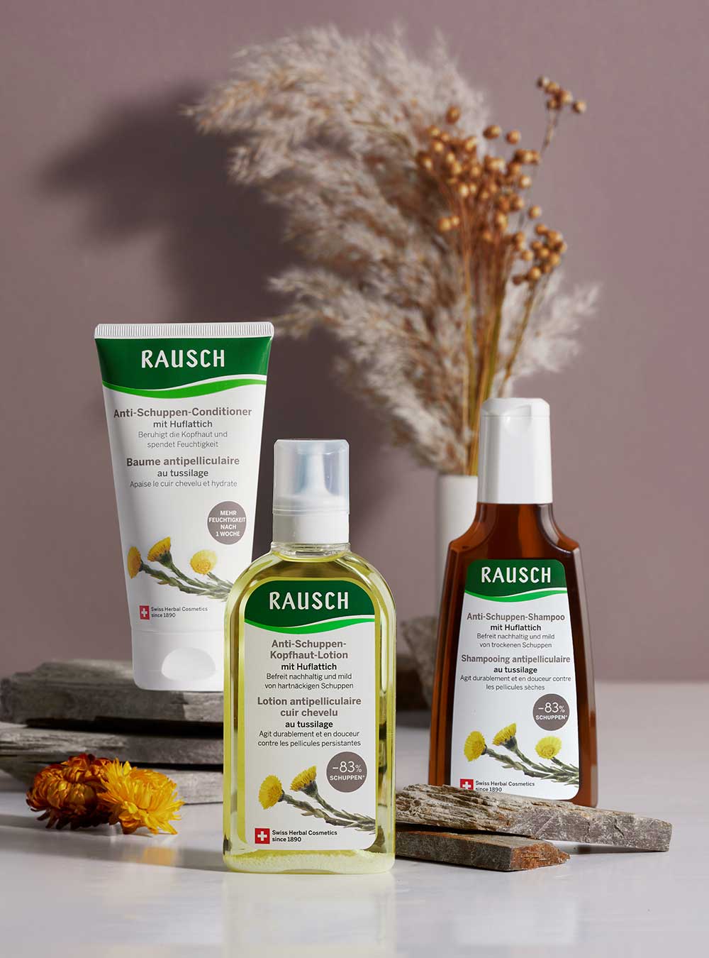 Three Rausch haircare products arranged on a tabletop with dried flowers and stone props: a white conditioner tube, a clear bottle of hair oil with pale yellow liquid, and an amber shampoo bottle. The products have green and white labels with botanical illustrations.