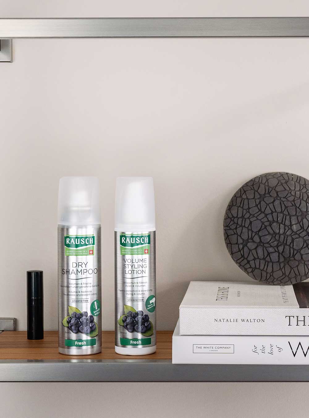 Two Rausch hair care products, Dry Shampoo and Volume Strengthening Lotion, standing on a wooden shelf next to books and a decorative object against a neutral wall.