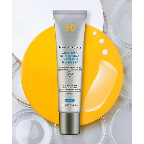 A tube of SkinCeuticals Advanced Brightening UV Defense Sunscreen Lotion SPF 50, presented on a bright yellow circular prop with white cosmetic droplets and clear glass lids in the background.